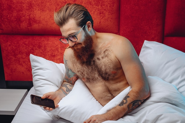 眼鏡をかけ、白いベッドに座って寝る前に電話を見ている男