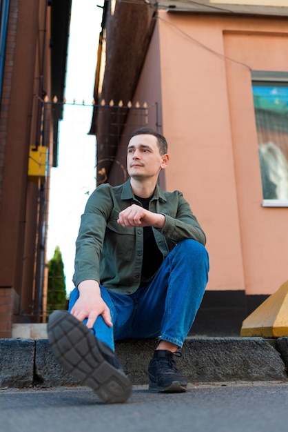 A man wearing a green shirt and jeans sitting on the sidewalk and looks towards