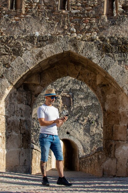 Man wearing hat and sunglasses standing in the doorway of an old stone castle while checking his cell phone