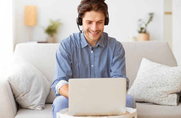 A man wearing headphones sits on a couch in his home smiling and working on a laptop