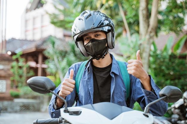 Man wearing helmet and mask with thumbs up on a motorcycle in the street background