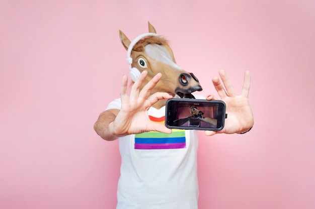 분홍색 배경이 있는 스튜디오에서 스마트폰으로 셀카를 찍는 동안 말 마스크를 쓴 남자