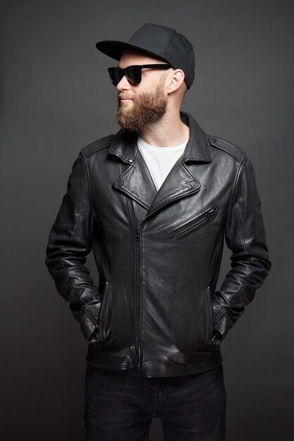 Man wearing leather biker jacket or asymmetric zip jacket with black cap, jeans and sunglasses. Handsome hipster man over gray background.