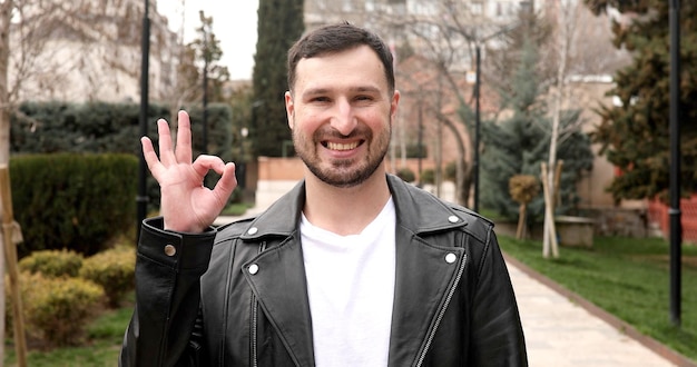 A man wearing a leather jacket and a white shirt shows an ok sign.