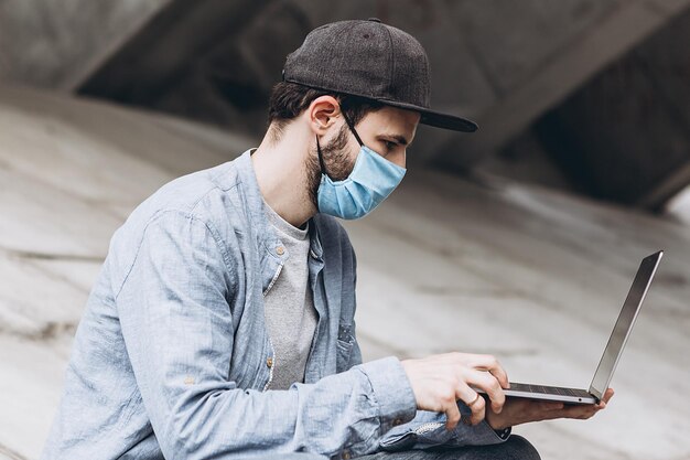 Foto uomo con maschera e abbigliamento casuale con il portatile in mano che lavora in remoto all'esterno