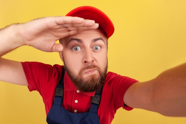 Man wearing overalls taking selfie looking far with hand over head having ridiculousexpression POV