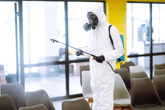 Man wearing protective suit disinfecting assembly hall with spray chemicals preventing coronavirus