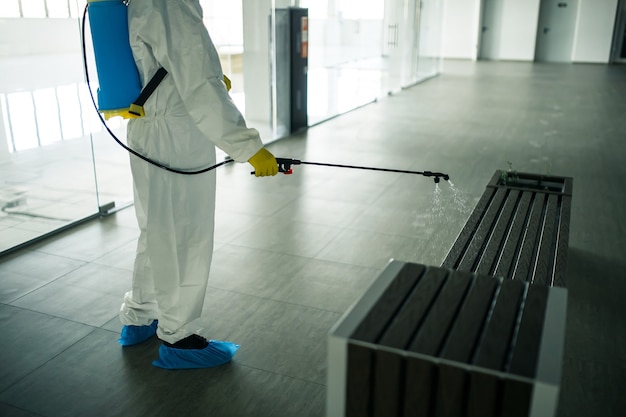 A man wearing protective suit is disinfecting a bench in an empty shopping mall with sanitizing spray. Cleaning up the public place to prevent covid spread. Healthcare precautions and safety concept.