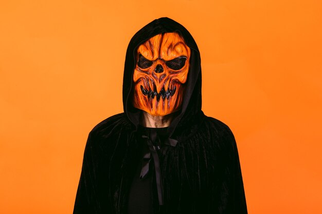 Photo man wearing pumpkin latex mask and hooded velvet cape, on orange background. halloween and days of the dead concept.