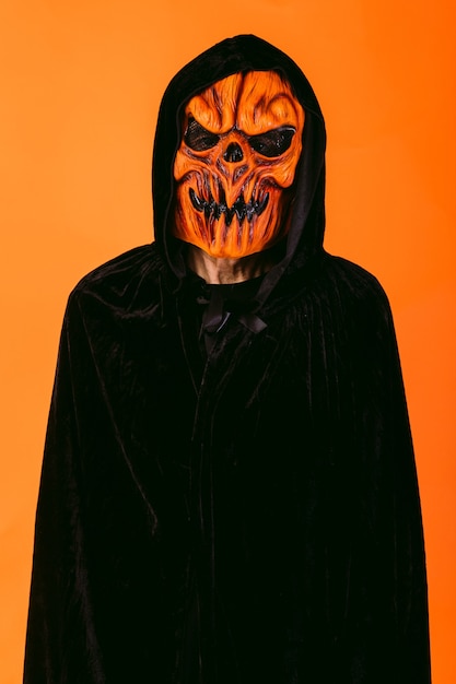 Man wearing pumpkin latex mask and hooded velvet cape, on orange background. Halloween and days of the dead concept.