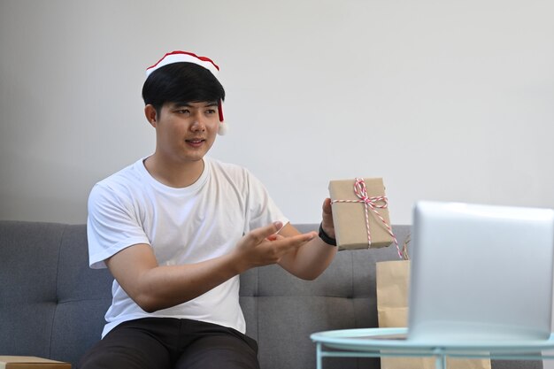 Man wearing red Santa hat  showing Christmas present during video call with his family.