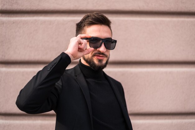 A man wearing sunglasses and a black turtleneck stands in front of a wall.