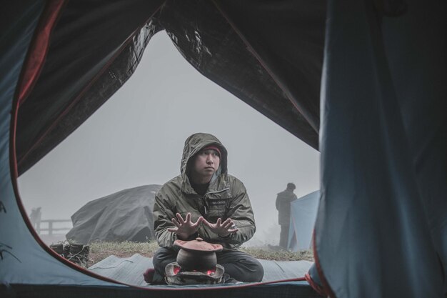 Photo man wearing warm clothing while sitting by camping stove seen through tent