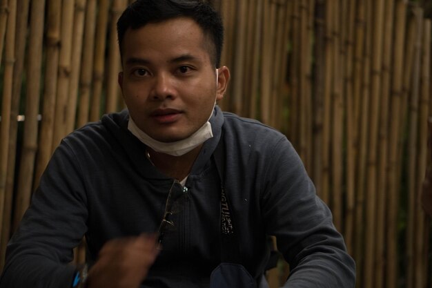 A man wearing a white mask sits at a table in front of bamboo poles.