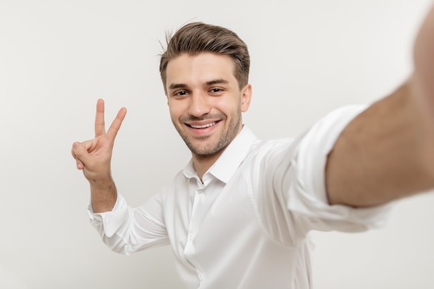 白いシャツを着た男が白い背景で隔離の2本の指の平和ジェスチャーを示す自分撮りを作る