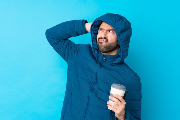 Photo man wearing winter jacket and holding a takeaway coffee over isolated blue wall having doubts