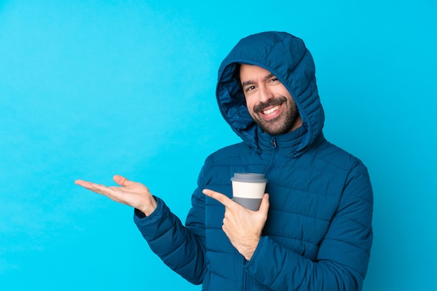 Человек, одетый в зимнюю куртку и проведение вынос кофе на изолированной синей стене, держа воображаемое copyspace на ладони, чтобы вставить объявление