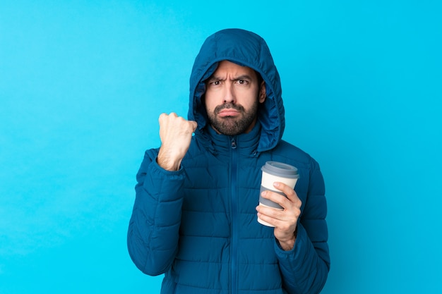 Man wearing winter jacket and holding a takeaway coffee over isolated blue wall with angry gesture