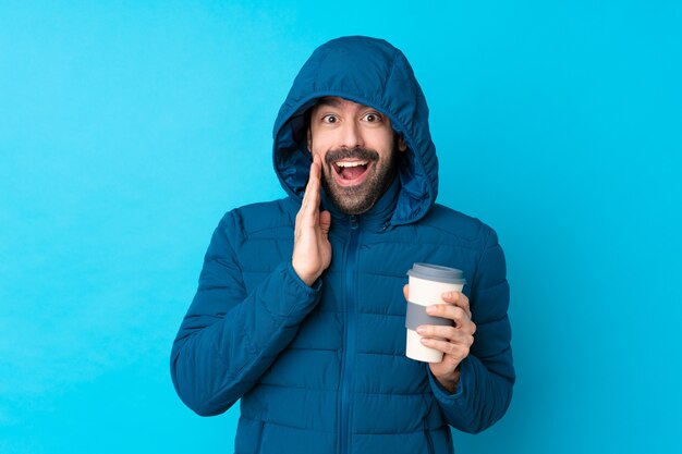 Man wearing winter jacket and holding a takeaway coffee over isolated blue wall with surprise and shocked facial expression