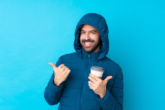 Man wearing winter jacket and holding a takeaway coffee over isolated blue wall with thumbs up gesture and smiling