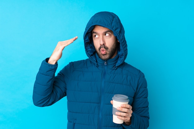 Man wearing winter jacket and holding a takeaway coffee over isolated blue wall with tired and sick expression