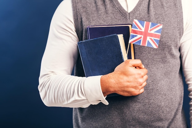 Man wears grey sweater vest holds english books and flag before dark blue studio background language learning concept