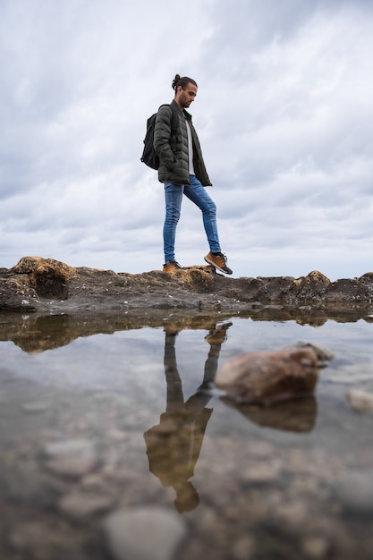 Man weerspiegeld in een plas in de zee. Op een bewolkte dag. Hij draagt een groene jas, een grijze trui en een spijkerbroek. Blank, met paardenstaart en rugzak op zijn rug.