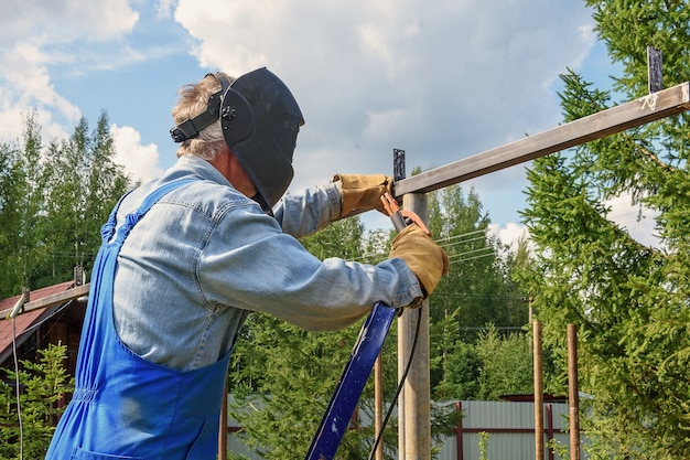 man welder in a welding mask, construction uniform and protective gloves cooks metal on a street construction site. construction of a pavilion, pergola near a country house on a summer day.