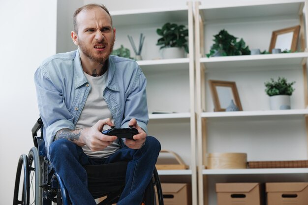 Photo a man in a wheelchair gamer plays games with a joystick in his hands at home anger and sadness from losing copy space with tattoos on his hands health concept man with disabilities real person