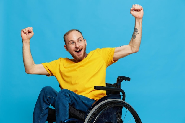 A man in a wheelchair happiness raised his hands up in a tshirt with tattoos on his hands sits on a blue studio background a full life a real person