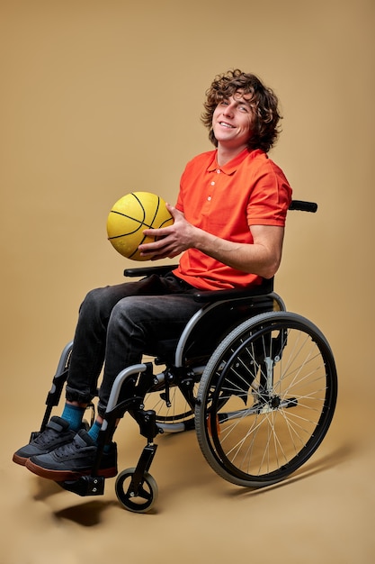 Man on wheelchair is playing a ball to strengthen muscles, holding yellow basketball ball. lifestyle of disability people, isolated beige background