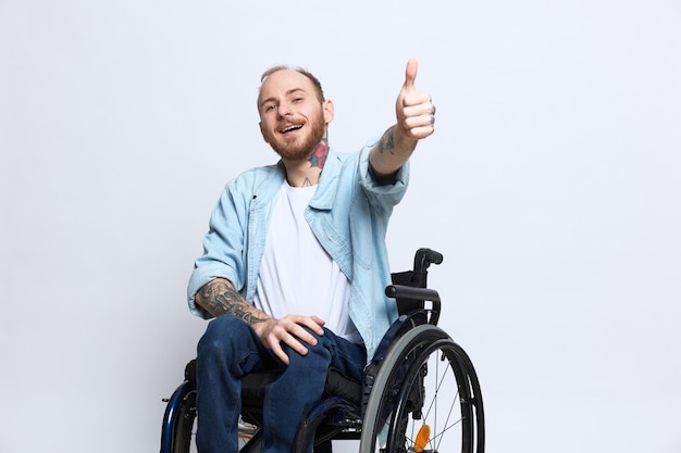車椅子に乗った男性がカメラを見て、手にタトゥーを入れて親指を立てて幸せを示し、灰色のスタジオの背景に座って健康コンセプトを持っている障害を持つ男性