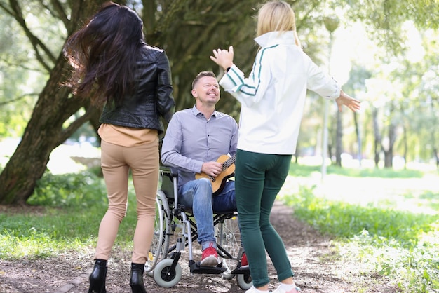 A man in a wheelchair plays the ukulele for women in the park closeup dances and songs in the