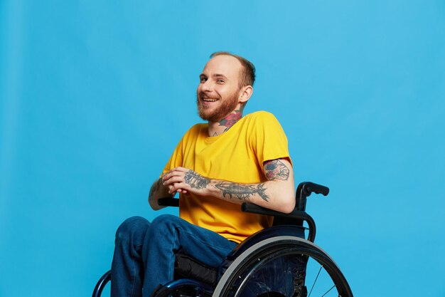 A man in a wheelchair smile looks at the camera in a tshirt with tattoos on his arms sits on a blue studio background a full life a real person