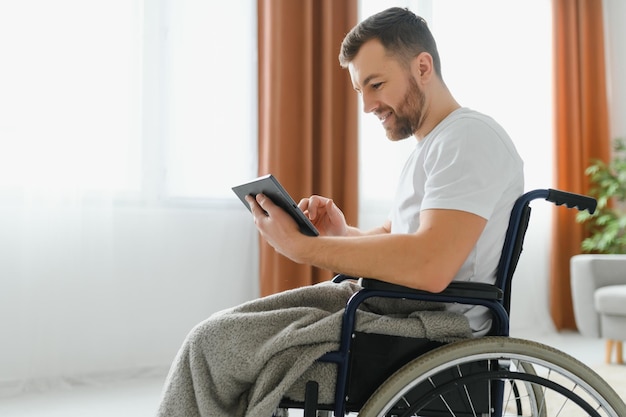 Man in wheelchair using tablet computer at home Positive retired male with physical disability browsing web on touch pad watching video online indoors