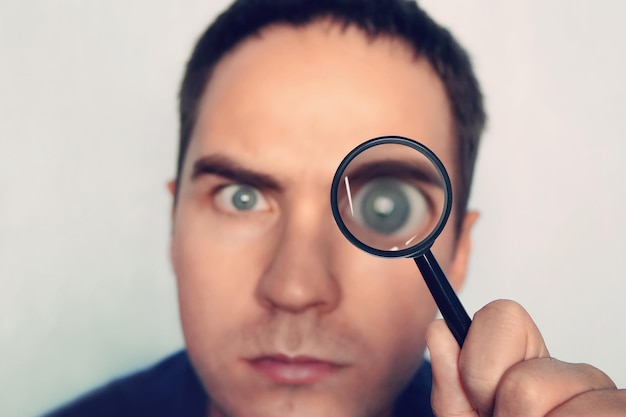 Man on white background curiously looks through the magnifying glass Male fuzzy eye magnified through the loule Person uses magnifier to look closely at some object Detective search for evidence