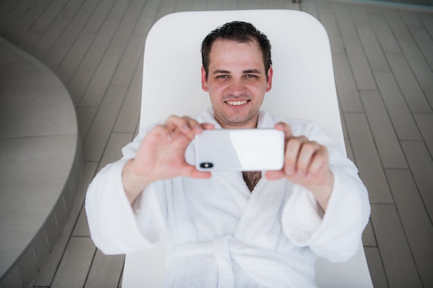 Un uomo con un accappatoio bianco giace a bordo piscina su una sedia a sdraio e guarda il nuovo costoso cellulare