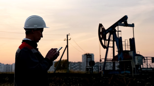 Человек в белом шлеме с телефоном на фоне качающейся нефтяной скважины и закатного неба