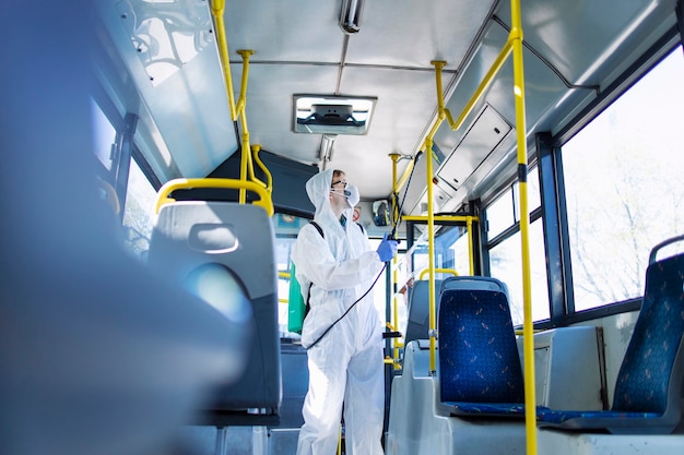 Man in white protection suit disinfecting handlebars of bus interior to stop spreading highly contagious corona virus