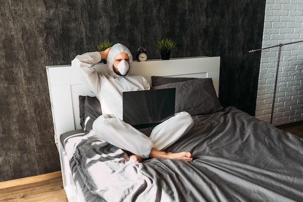 Man in white protective costume with hood and face mask at home on the bed with laptop