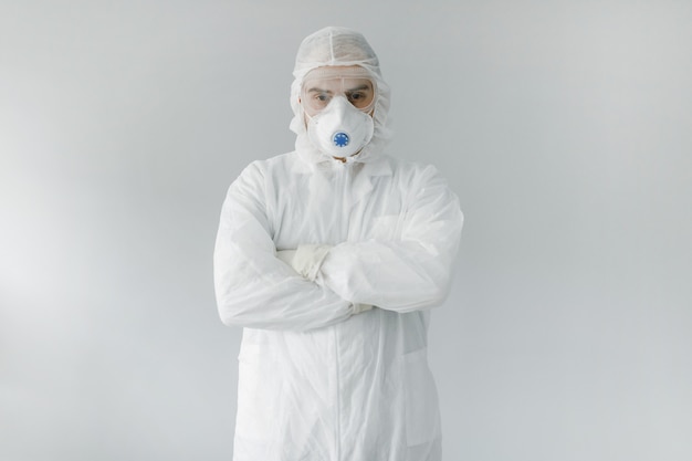 Man in white protective suit, mask, glasses and gloves is coughing on white background, coronavirus pandemic threat.