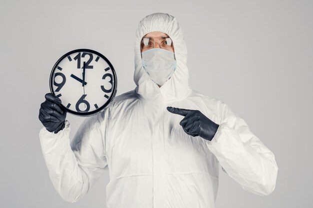 A man in a white protective suit a mask on a white background with a watch in his hands An epidemic pandemic is a new rapidly spreading virus a medical concept