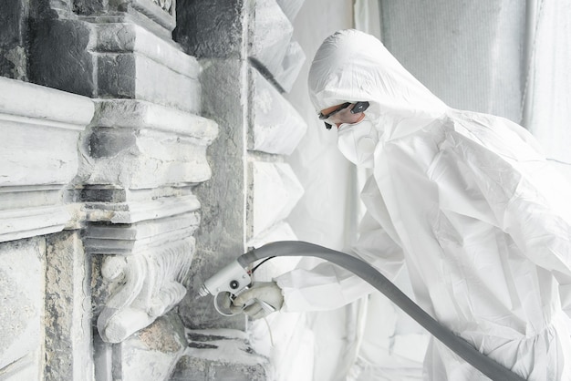Photo man in a white protective uniform cleans stone carved sculpture from the dirt and concrete with a sandblasting machine. restoration of stone sculpture. a jet of sand under high pressure.