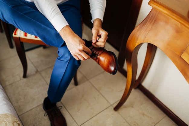 Photo man in a white shirt and blue trousers ties his boots sitting on a chair closeup