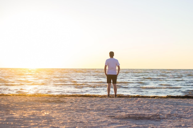 A man in a white shirt and brown shorts standing on empty beach at sunrise