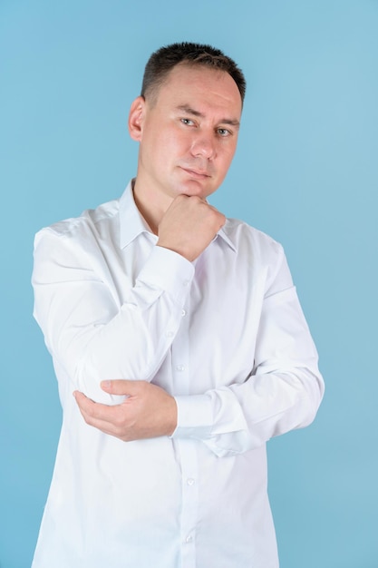 A man in a white shirt holds his hand to his chin and looks at the camera standing on a blue background