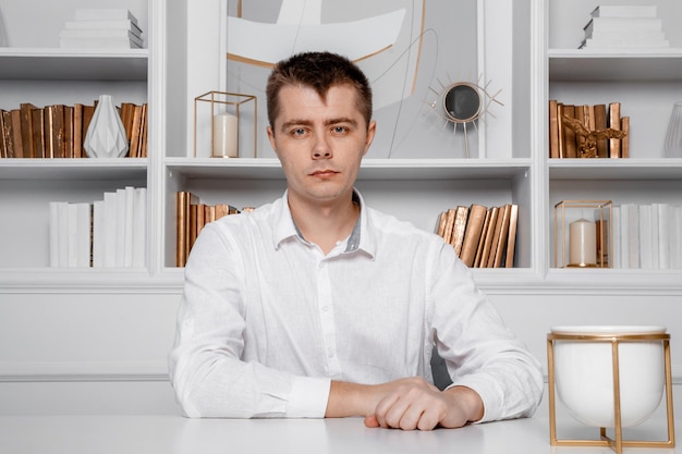 A man in a white shirt is sitting at a white table. Young business man on a desk. Attractive businessman sitting at table. Front view of a business man working. Portrait.