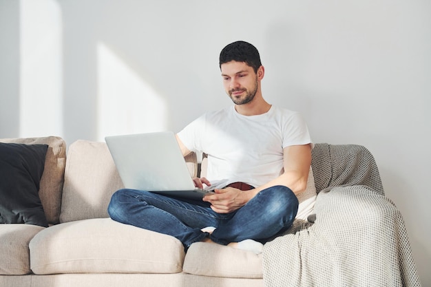 Photo man in white shirt and jeans sitting on bed with laptop indoors