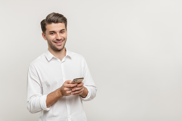Photo man in white shirt standing holding mobile phone and smiling at camera isolated on white background
