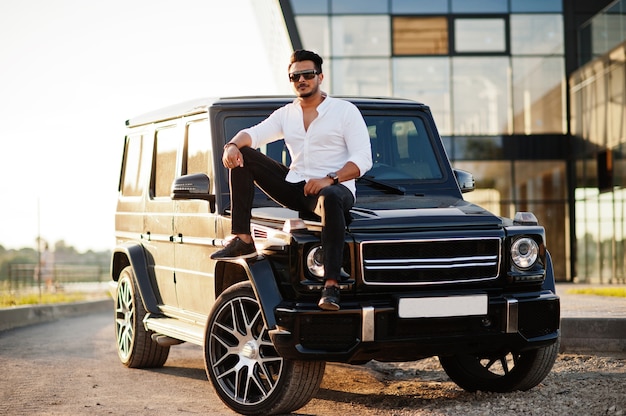 Man in white shirt and sunglasses posing near black suv car